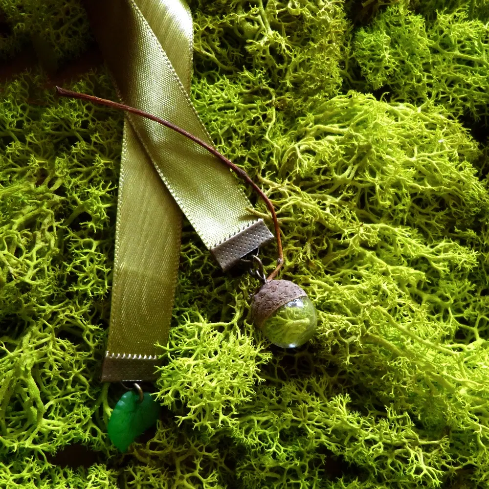 Grünes Band-Lesezeichen, an dem ein Eichelhütchen hängt, in das ein Glasanhänger mit eingeschlossenem Moos eingefasst ist.