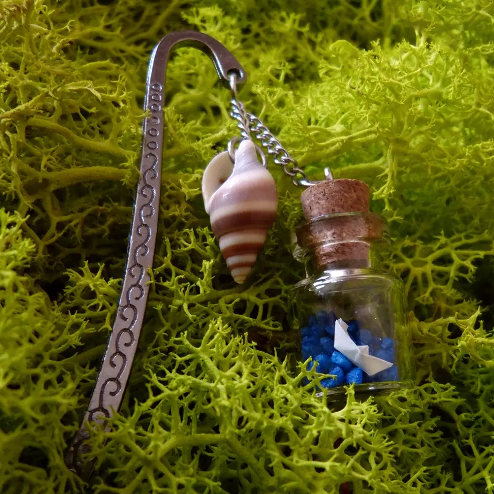 Metall-Lesezeichen mit graviertem Muster. An einer feinen Kette hängen eine Muschel und ein Glasfläschchen mit blauem Kies und einem gefalteten Papierschiff.